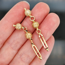 Load image into Gallery viewer, A pair of engraved bead and bar drop earrings in yellow gold, held in a hand