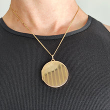 Load image into Gallery viewer, A circular yellow gold locket with pinstripe engraving, with a chain, modelled by a woman wearing a black top
