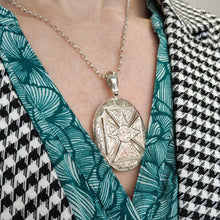 Load image into Gallery viewer, An intricate engraved oval locket pendant in silver, modelled with a chain by a woman wearing a green patterned top and houndstooth print jacket