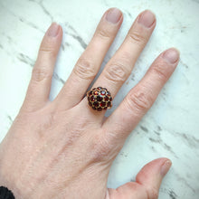 Load image into Gallery viewer, A round garnet domed cluster ring in yellow gold, modelled on a middle finger, against a marbled background