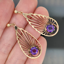 Load image into Gallery viewer, A pair of pear shaped fan design drop earrings with amethysts in yellow gold, held in a hand
