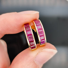 Load image into Gallery viewer, A pair of yellow gold hoop earrings channel set with square cut rubies, held in a hand
