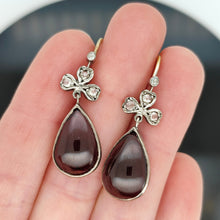 Load image into Gallery viewer, A pair of pear shaped garnet and rose cut diamond earrings in gold and silver, held in a hand

