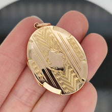 Load image into Gallery viewer, An oval yellow gold locket pendant with engraving, held in a hand
