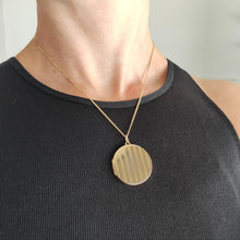 Load image into Gallery viewer, A circular yellow gold locket with pinstripe engraving, with a chain, modelled by a woman wearing a black top
