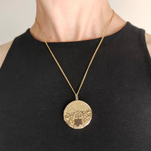 Load image into Gallery viewer, A circular yellow gold engraved locket pendant, with a chain, modelled by a woman wearing a black top
