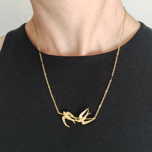 Load image into Gallery viewer, A yellow gold pendant necklace featuring two swallow birds in flight, modelled by a woman wearing a black top
