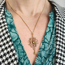 Load image into Gallery viewer, A peridot and pearl Art Nouveau style pendant in yellow gold, on a chain, modelled by a woman wearing a green patterned shirt and houndstooth jacket
