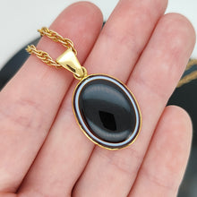 Load image into Gallery viewer, An oval banded agate pendant rubover set in yellow gold, with a yellow gold chain, held in a hand
