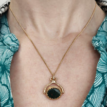 Load image into Gallery viewer, An oval spinner fob pendant in yellow gold with bloodstone, on a chain, modelled by a woman wearing a green patterned top
